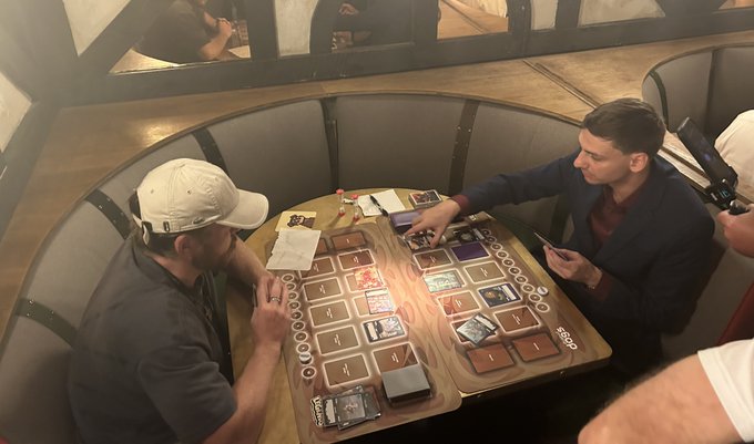 Two men seated across from each other at a circular wooden table, engaged in a tabletop card game with playing cards and game pieces.