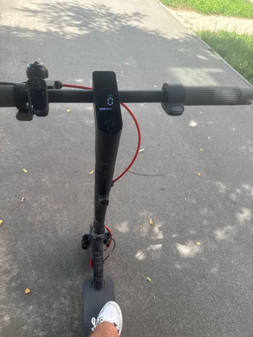 A first-person view of someone riding an electric scooter on a paved path, with the digital display showing '0' and white sneakers visible.