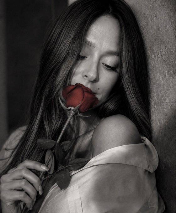 A black-and-white portrait of a woman with long hair holding a red rose near her face, leaning against a wall.