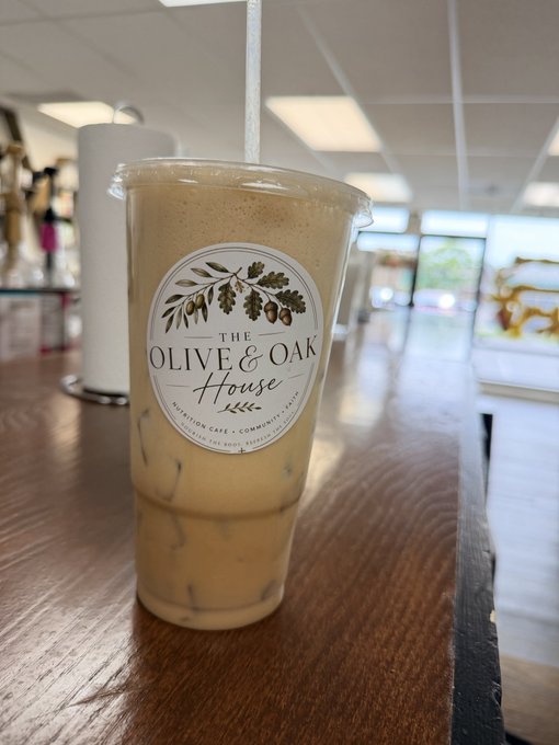 A clear plastic cup containing an iced coffee drink sits on a wooden counter, branded with 'The Olive & Oak House' logo featuring an olive branch design.
