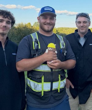 Three men stand outdoors near a construction or worksite, with the central man wearing a blue cap and safety vest holding a Lego minifigure, while the two men on either side wear dark jackets.