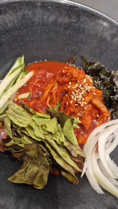 A close-up of a bowl of food containing red sauce, vegetables, greens, noodles, seaweed, and sesame seeds.