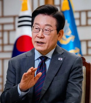 South Korean politician Lee Jae-myung is shown speaking, wearing a gray suit and striped tie, with a South Korean flag and a presidential standard in the background.