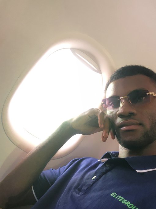 A young man wearing sunglasses and a navy blue 'ELITEGROU' polo shirt poses for a selfie beside an airplane window with bright sunlight streaming through.