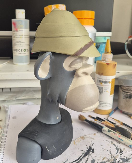 A sculpture in progress showing a gray monkey head wearing a tan pith helmet with a brown band, sitting on a work surface surrounded by paint bottles and brushes.