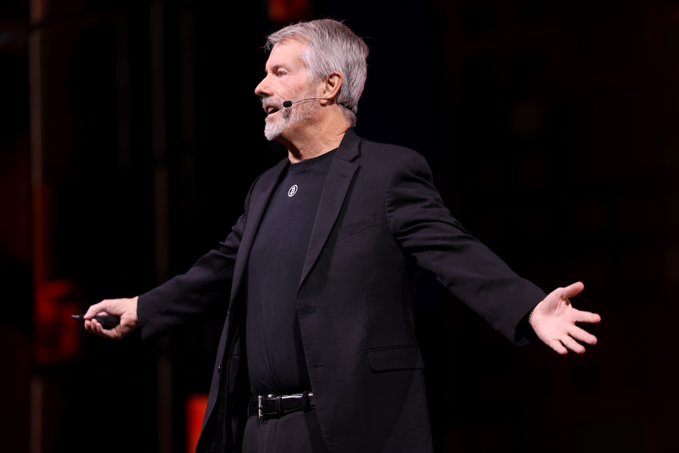 Michael Saylor, founder of MicroStrategy and a prominent Bitcoin advocate, speaks on stage wearing a headset microphone and black blazer with arms outstretched in an expressive gesture. The image captures him during what appears to be a public address consistent with his recent bullish remarks declaring Bitcoin's dominance over competing cryptocurrencies like Solana and Ethereum.