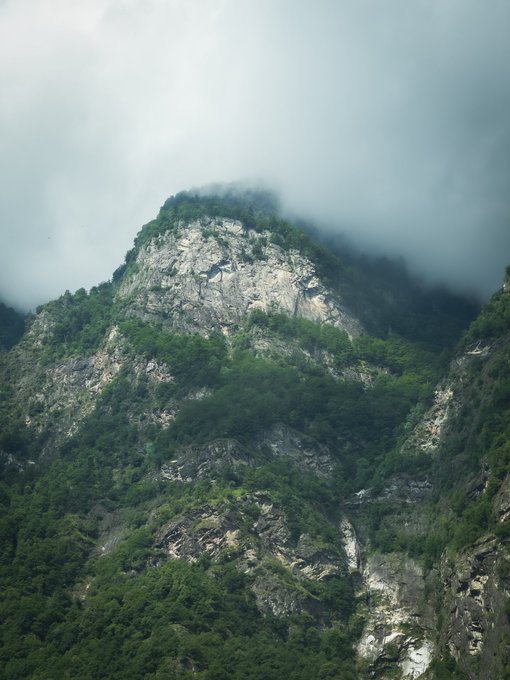 The image shows a mountain covered with trees, rocks, and a large rock formation at its peak. The sky above the mountains has clouds, creating an atmospheric scene. The photo appears to be taken from a high vantage point, providing a bird's eye view of the landscape below.