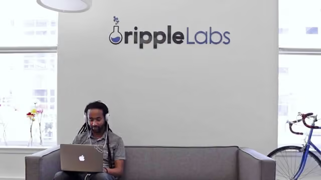 A man wearing headphones works on a laptop while seated on a couch in the Ripple Labs office, with the company's logo prominently displayed on the wall behind him. The image connects to the post about Ripple's XRP Ledger upgrading its infrastructure to target the RWA (Real World Asset) sector, aiming to attract $530 million in assets from Wall Street funds as the company seeks new growth drivers beyond its traditional cross-border payment business.
