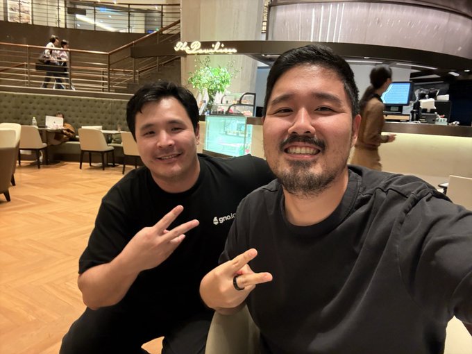 Two smiling Asian men pose for a selfie in a modern cafe or restaurant, both wearing black shirts with one making a peace sign with his hand.