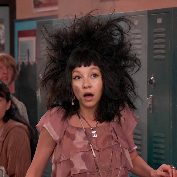 A still image of actress/singer Jenna Ortega with dramatically voluminous, frizzy black hair, wearing a pink ruffled top, appearing to be from a TV show scene.