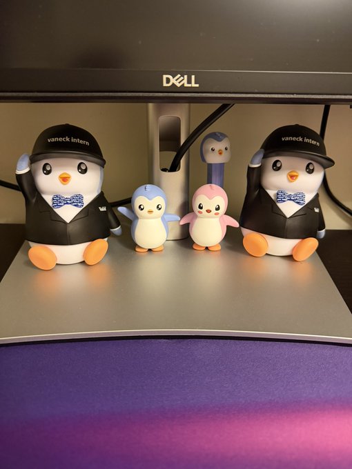 Five penguin figurines displayed on a Dell monitor stand, including two large ones wearing black 'vaneck intern' suits and bow ties, and three smaller colorful penguins (blue, pink, and dark blue) in the middle.