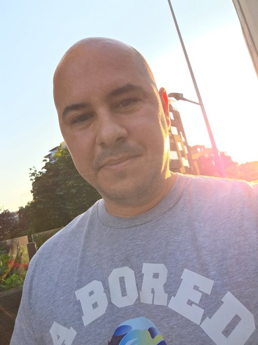 A bald man takes a selfie wearing a gray t-shirt that reads "BORED" with a small Bored Ape character visible, captured against a sunset sky as he wraps up his time in Sofia before continuing his drive.