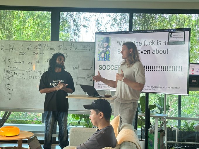 The image shows two presenters standing in front of a whiteboard and a large screen displaying "SOCCER" with the phrase "What the fuck is the B...even about" visible, while addressing a seated audience. This matches the post's description of a World Cup track prebriefing where speakers broke down building on live World Cup data via the TxLINE API on Solana, with MagicBlock representatives presenting.