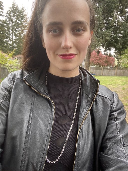 A selfie of a young woman wearing a black leather jacket and silver chain, standing outside with trees and a fence in the background.