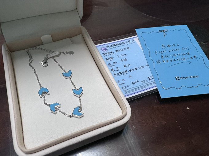 The image features a silver bracelet with blue accents placed inside a white jewelry box. The bracelet is positioned next to an envelope or card that has Chinese characters written on it, suggesting that this could be a gift from China. The bracelet appears to be new and unused, as indicated by its pristine condition.