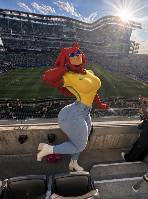 A cosplayer with red hair, sunglasses, a yellow Nike top, and blue jeans poses on stadium seats overlooking a soccer field.