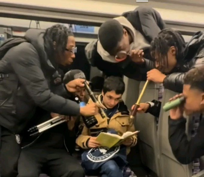 A group of young men crowd together on public transit, peering closely at a small green book being held by the person seated in the center.