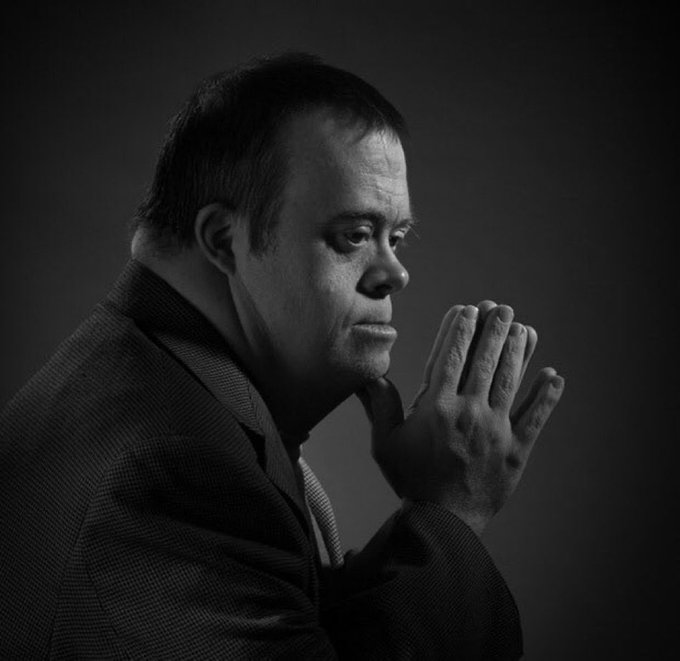 A black and white portrait photograph of a man in a suit and vest with his hands clasped together in a contemplative prayer-like pose against a dark background.
