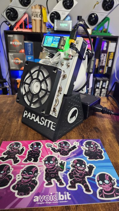 A close-up photograph of a custom computer build featuring a large fan and the word 'PARASITE' on the base, sitting on a desk with a sticker sheet showing various cartoon characters and the text 'avoidbit' in the foreground.