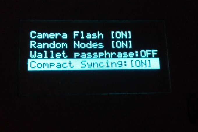 Close-up photo of an electronic device's LCD settings menu displaying options: "Camera Flash [ON]", "Random Nodes [ON]", "Wallet Passphrase: OFF", and the highlighted "Compact Syncing: [ON]".
