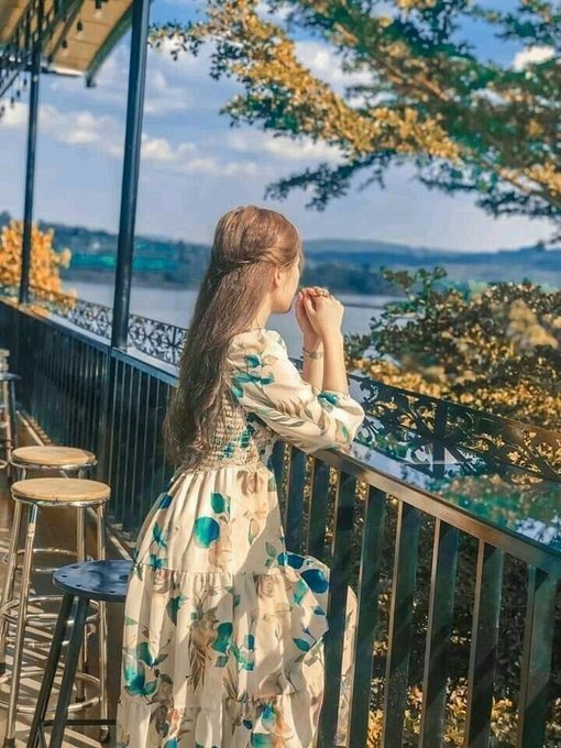 A woman with long brown hair wearing a cream-colored floral dress stands on a balcony with wrought iron details, looking out at a lake and distant hills on a sunny day.