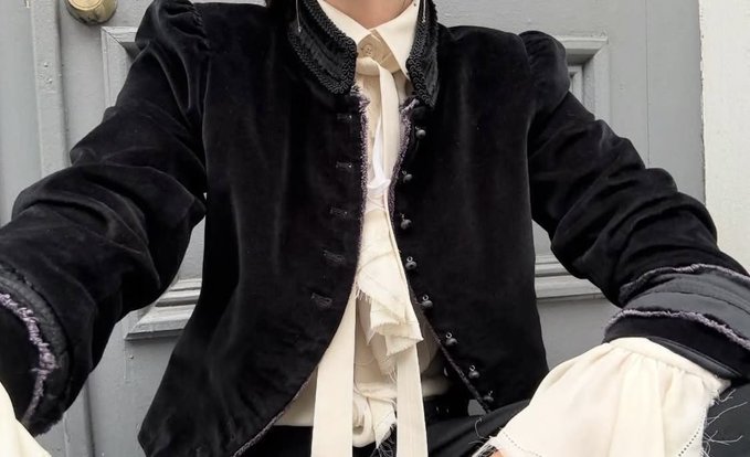 A close-up photo of a person wearing a dark, velvet-like jacket with frayed edges and decorative buttons over a cream-colored ruffled blouse, standing in front of a gray paneled door.