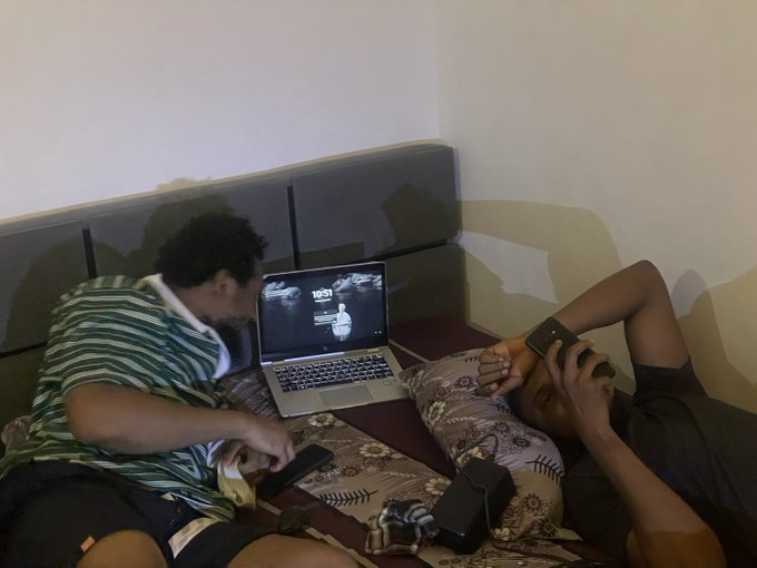 Two young men relax on a bed; one sits up watching a laptop screen while the other lies down looking at a smartphone.