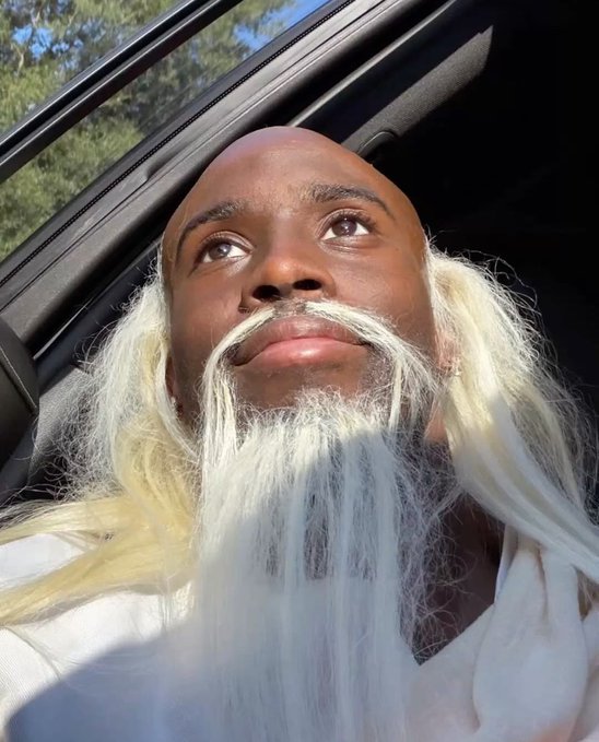 A bald Black man with a long, flowing white beard sits in a car, looking upward in a dramatic low-angle selfie.