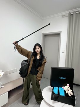 A young woman with long dark hair wearing a tan jacket and olive green pants stands in a room holding a black selfie stick raised above her head, posing next to a PlayStation 5 console displaying a game on a small table.