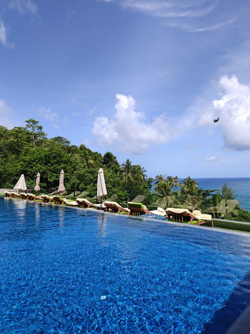 A scenic view of an infinity pool overlooking a tropical beach with palm trees, ocean, and a partly cloudy blue sky.