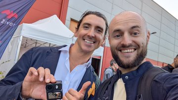 The image shows two men standing next to each other outside a building with a red wall. One of them is holding up a small camera or camcorder, possibly taking a photo or recording something interesting happening around them. The man on the left has his hand raised in front of him and is smiling at the camera while the man on the right looks towards the camera. There are other people present in the background as well, suggesting that they might be attending an event together.