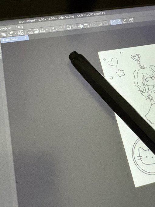 A black stylus pen resting on a tablet displaying the Clip Studio Paint EX interface, showing a sketch of a girl with hearts and a cat illustration.