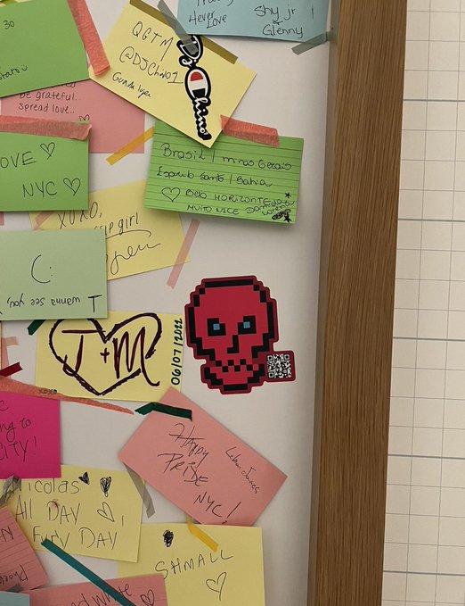 A white board or door is covered in colorful sticky notes with handwritten messages like 'NYC', 'STANNYC', and '090109', along with a pixelated red skull sticker and a heart with 'J+M'.
