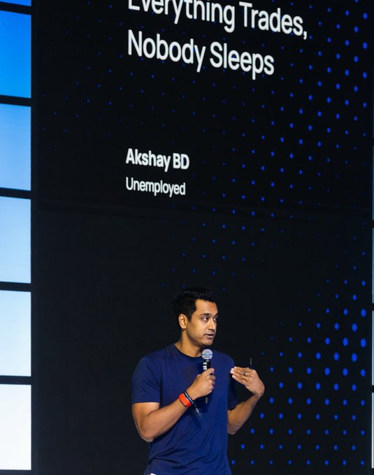 A man named Akshay BD (listed as 'Unemployed') speaks into a microphone on stage with the presentation slide titled 'Everything Trades, Nobody Sleeps' displayed behind him.