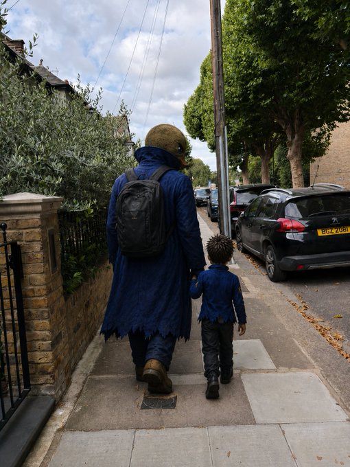 A person in a tattered blue cloak and backpack wearing a large round helmet walks hand-in-hand down a sidewalk with a small child in a blue shirt, alongside parked cars including a black Peugeot with license plate BGZ 2081.
