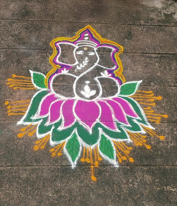 A colorful rangoli design on concrete featuring Ganesha seated on a pink lotus with green leaves and yellow accents.