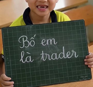 A young child holding up a black cutting mat with handwritten chalk text reading 'Bố em là trader' (My dad is a trader) in Vietnamese.