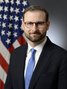 The image shows a man with glasses, a beard, and a dark suit with a blue tie, standing in front of an American flag, appearing to be an official portrait. Based on the accompanying post, this is Patrick Witt, the crypto policy chief referenced in connection with the CLARITY Act negotiations before his upcoming military leave.