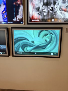 A gallery wall display featuring digital screens in black frames, with the prominent lower screen showing a swirling teal abstract graphic labeled 'SWIRL 01'. Other screens display various artwork and viewer counts.