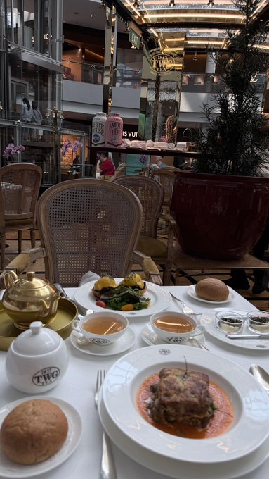 An elegant dining setup at a TWG tea location, featuring a gold teapot, a teacup, bread rolls, a vegetable dish, and a main course plated on a white dish.