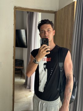 A man takes a mirror selfie in a sleeveless black shirt with graphic prints, accessorized with a watch and a shoulder bag, in a bedroom setting. The casual, confident vibe of the photo pairs with the giveaway post, projecting a relaxed and approachable energy befitting someone sharing good fortune with their followers.