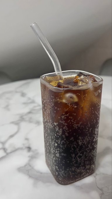 A tall glass filled with iced dark soda and bubbles sits on a marble surface with a glass straw inserted.