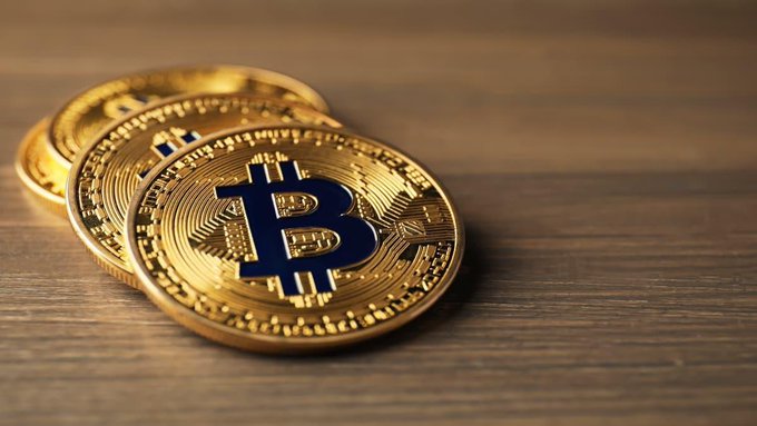 A close-up of two physical Bitcoin tokens with the recognizable blue 'B' logo resting on a dark wooden surface, showcasing the detailed circuit-like engravings on the gold-colored coins.