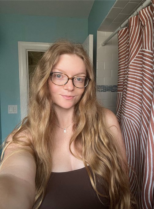 A selfie of a woman with long wavy blonde hair and glasses, wearing a dark tank top, standing in a bathroom with teal walls and a striped shower curtain.