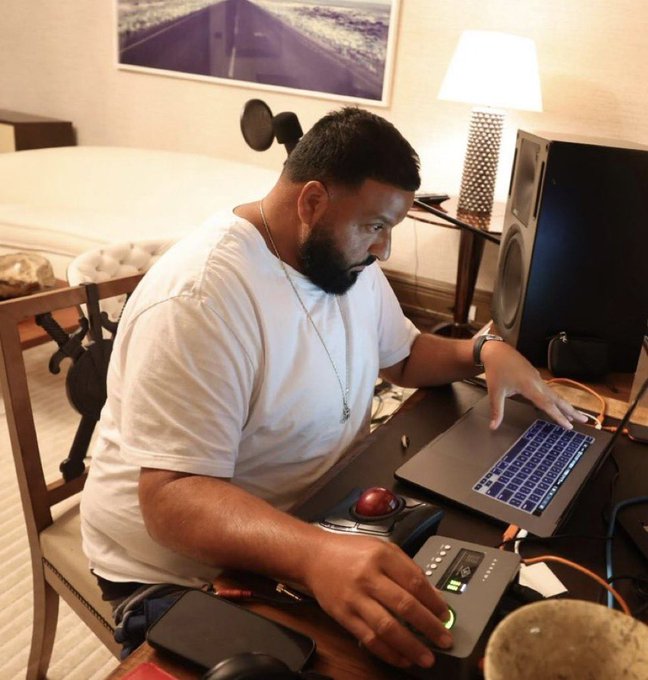 A man wearing a white t-shirt works on a laptop and audio equipment in what appears to be a hotel room.