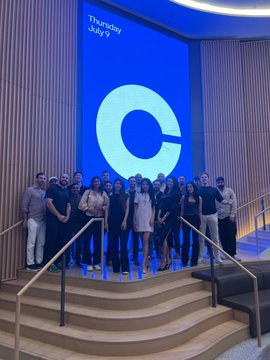 A group of people posing together on a stage in front of a large screen displaying a blue 'C' logo with text reading 'Thursday July 9'.