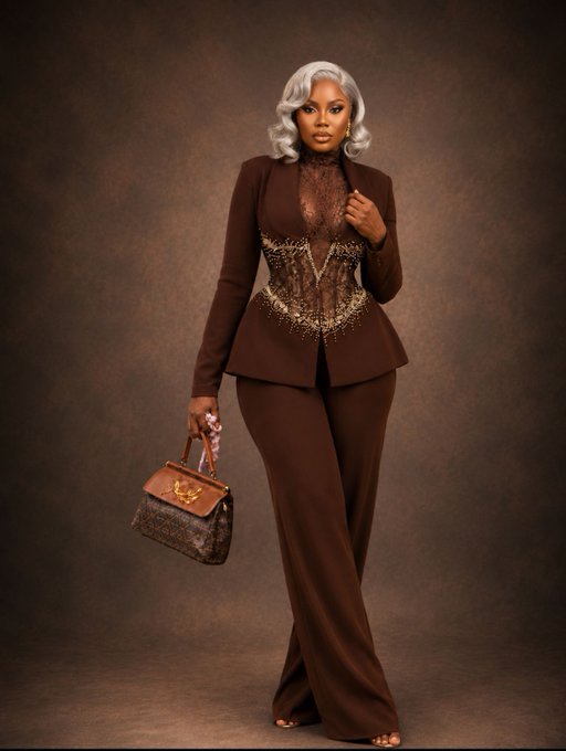 A fashion editorial photo of a model with a silver bob wig wearing a brown tailored jacket with a sheer, embellished bodice and wide-leg trousers, holding a brown monogram handbag.