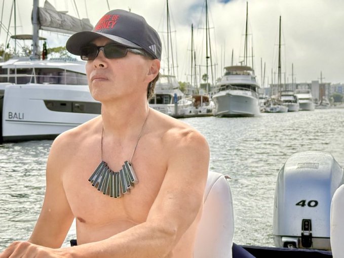 A shirtless man wearing a black 'Pirates of the Caribbean' Mickey Mouse cap, sunglasses, and a silver pendant necklace, sitting on a boat with the number 40 visible, with yachts including one named 'BALI' in the harbor behind him.