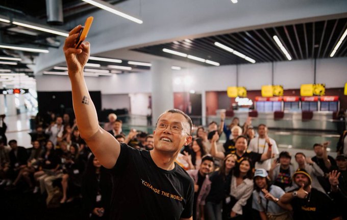 A man in a black t-shirt reading 'CHANGE THE WORLD' takes a selfie with a large crowd of fans behind him, appearing to be at a public event or gathering.