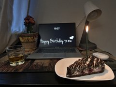 A birthday setup on a dark table features a lit candle, a slice of chocolate cake on a white plate, a glass of water, and a laptop displaying 'Happy Birthday to you'.